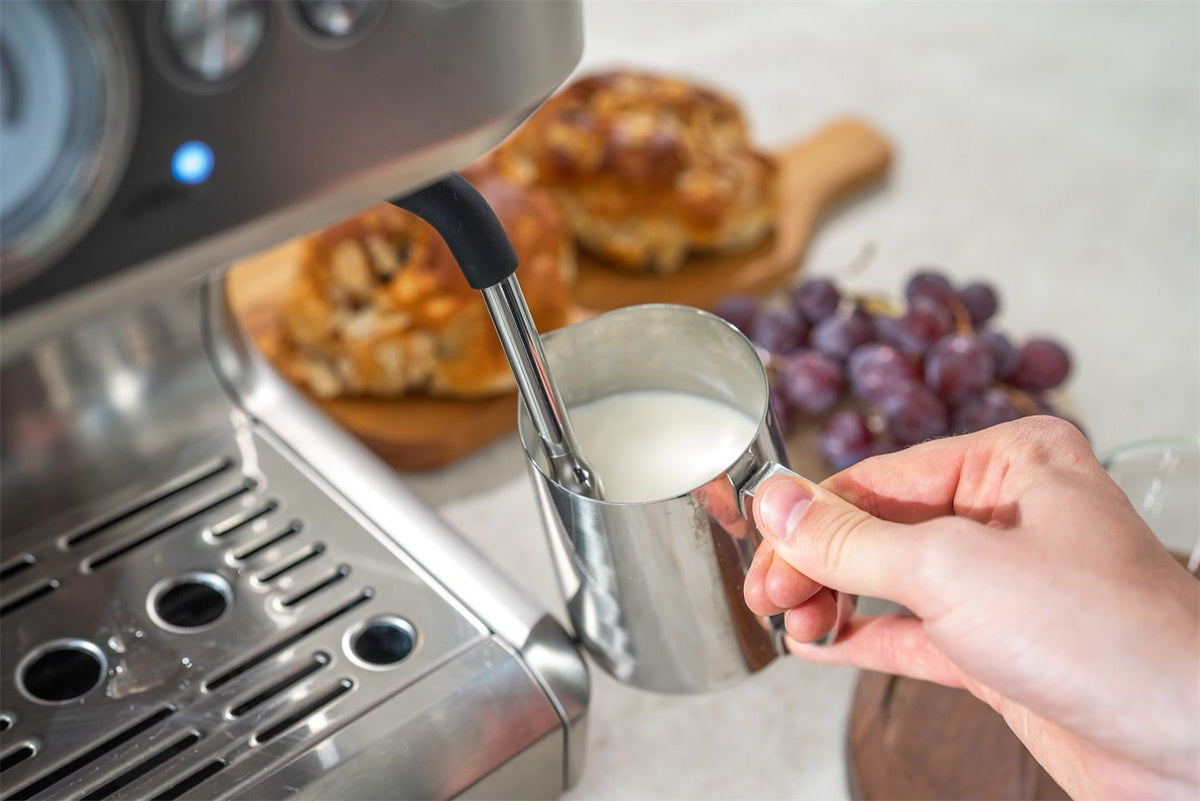 En hand håller en stålkanna med mjölk under ångröret på en espressomaskin i stål, med bröd och vindruvor i bakgrunden