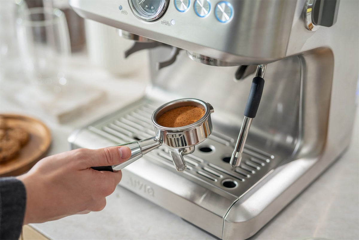 En hand håller ett portafilter med tampat kaffe framför en espressomaskin i stål