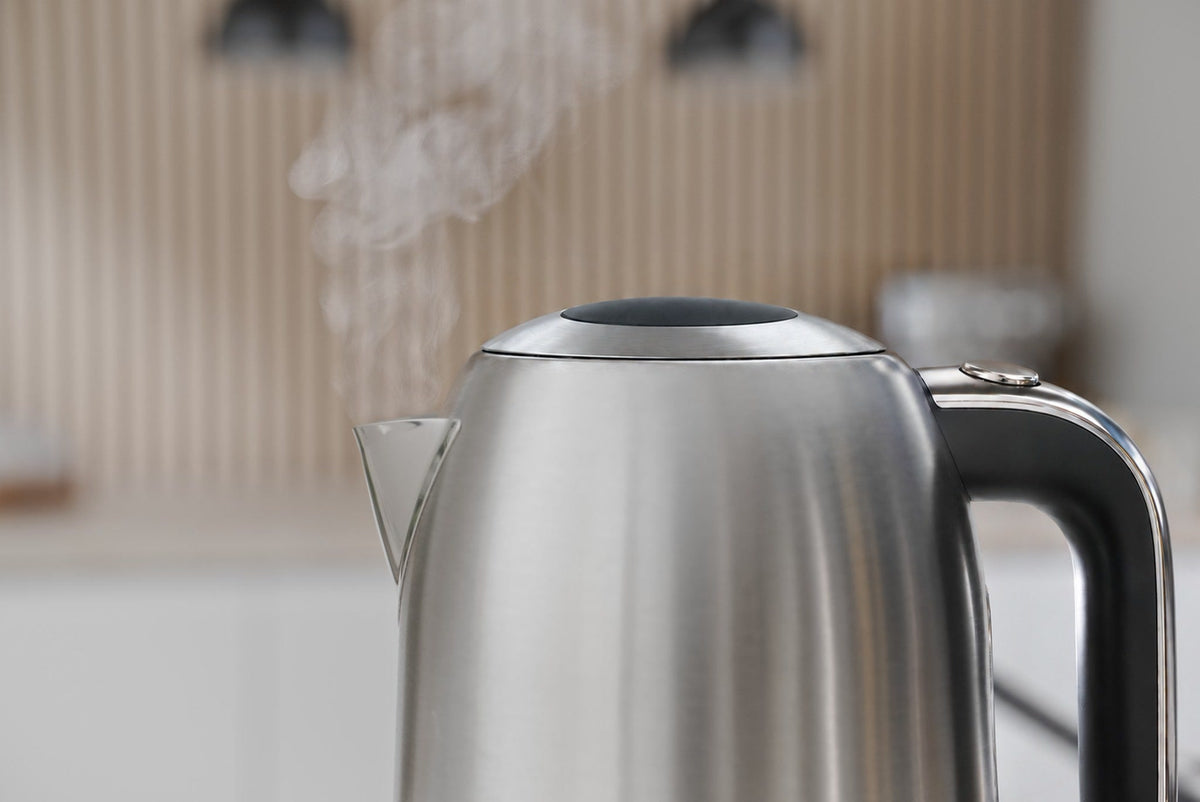 Vattenkokare i stål med ånga ur pipen framför en ljus vägg med lodräta träribbor