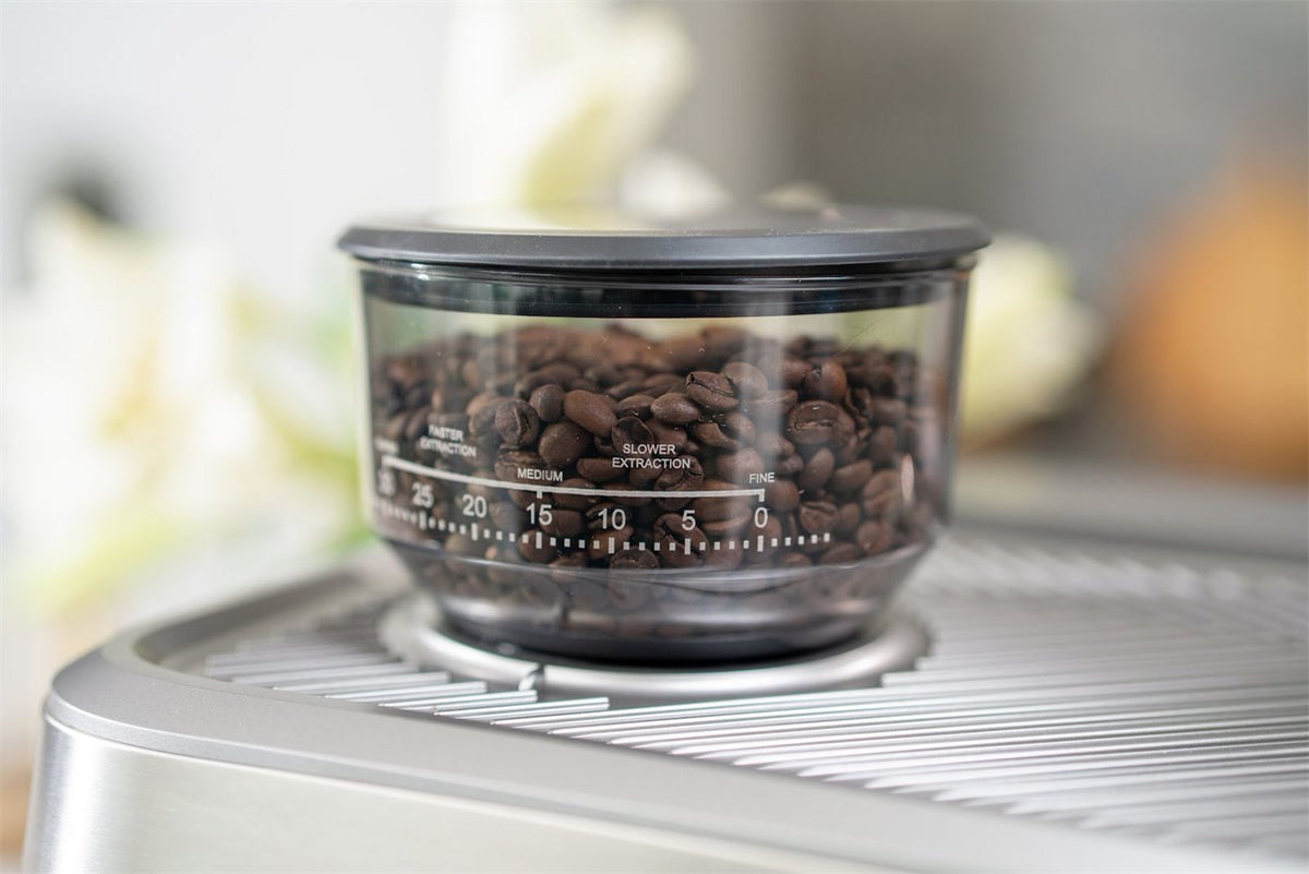Bönbehållare med kaffebönor och en skala från fin till grov ovanpå en espressomaskin i stål
