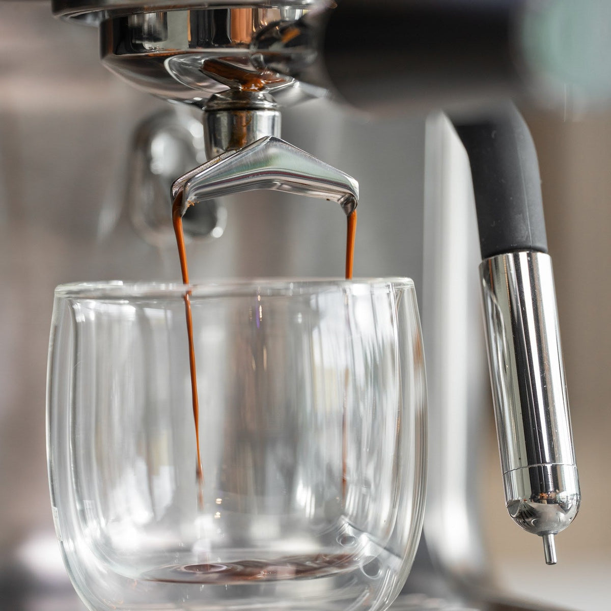 Espresso rinner i två tunna strålar från portafiltrets pip ner i ett dubbelväggigt glas. Till höger hänger ångröret.